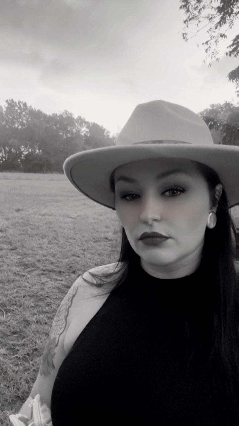 Black-and-white photo of Fawn in a wide-brimmed hat standing in an open East Texas field with trees behind her